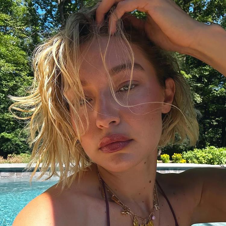 A imagem mostra Gigi Hadid com cabelo loiro curto, levemente bagunçado, posando ao ar livre. Ela usa um biquíni escuro e colares delicados. Ao fundo, há uma piscina azul clara e vegetação densa. A luz do sol ilumina seu rosto.