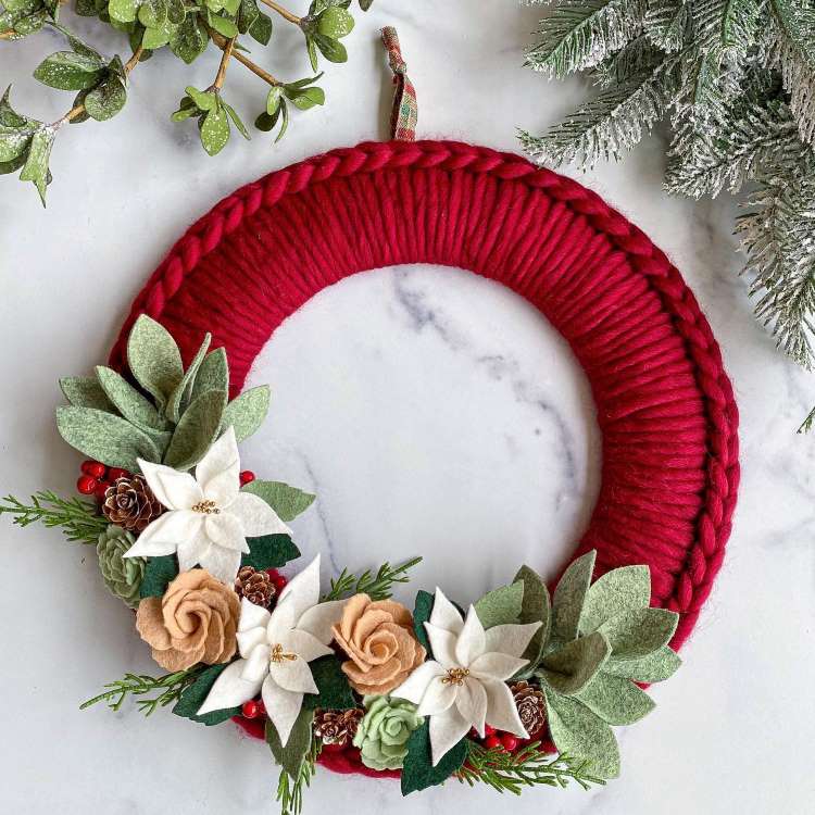 Guirlanda de Natal com base de tecido vermelho trançado. Decorada com flores de feltro em tons de branco e bege, folhas verdes, pinhas pequenas e bagas vermelhas. Situada em superfície de mármore com ramos de plantas ao redor, criando um visual festivo.