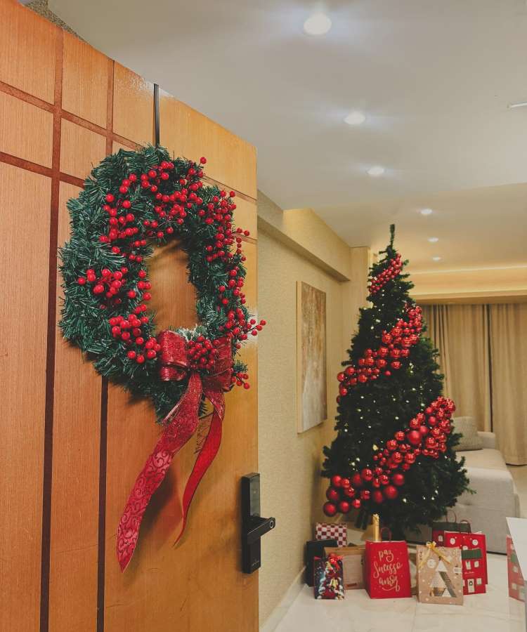 Enfeite de Natal de porta feito de ramos verdes com bagas vermelhas. Um laço vermelho elegante adorna a parte inferior. Ao fundo, uma árvore de Natal decorada com bolas vermelhas e presentes envoltos em papéis festivos, complementando o ambiente acolhedor.