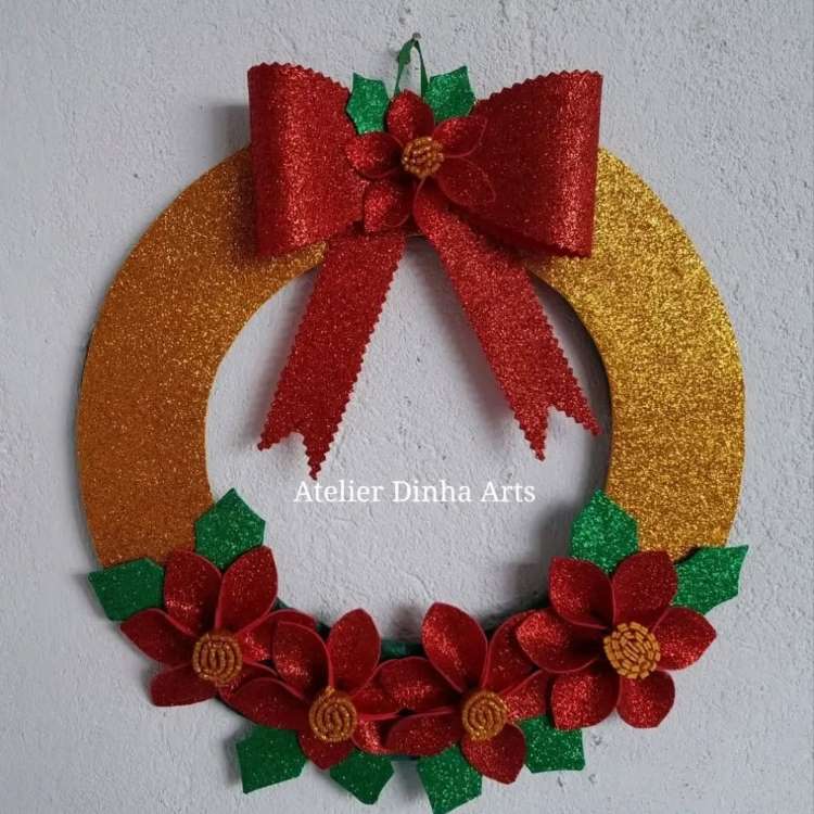 Guirlanda dourada com um laço vermelho brilhante no topo. Decorada com flores vermelhas com centros amarelos e folhas verdes embaixo. O design é elegante e festivo, perfeito para a decoração natalina, destacando-se sobre uma parede clara.