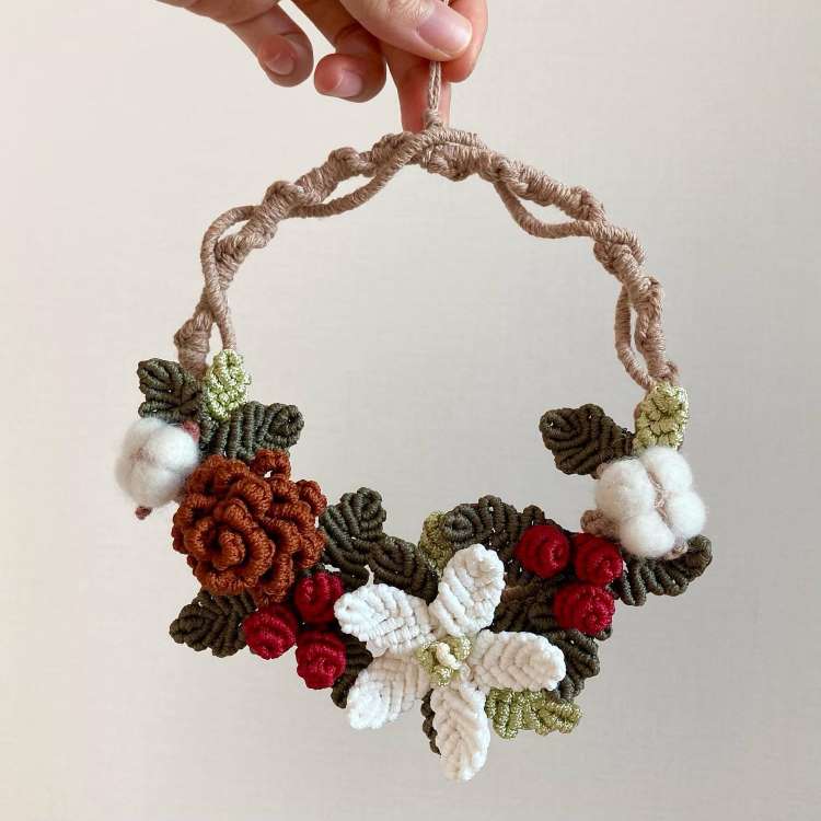 Um enfeite de Natal de porta artesanal feito de macramê. Decorado com flores brancas, vermelhas e marrom, além de folhas verdes e detalhes felpudos brancos. O design é rústico e delicado, ideal para adicionar charme natalino à decoração.