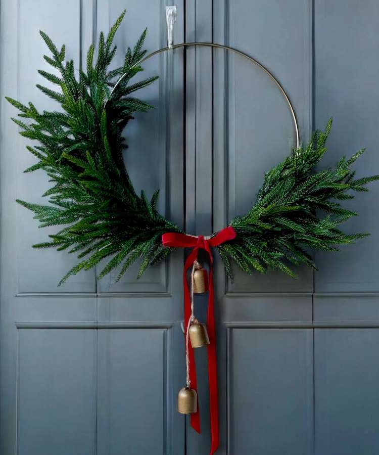 Enfeite de Natal de porta minimalista com estrutura metálica circular. Decorado com ramos verdes concentrados na parte inferior. Um laço vermelho elegante segura três sinos dourados pendentes. O fundo é uma porta azul escura, criando um contraste sofisticado e festivo.