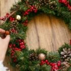 Enfeite de Natal de porta feito com galhos verdes, decorado com pinhas, bagas vermelhas e bolas douradas. Uma mão ajusta um detalhe na composição, sobre uma superfície de madeira clara, criando um toque caseiro e natural na decoração natalina.