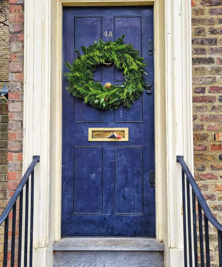 Enfeite de Natal de porta feito com ramos verdes naturais adornando uma porta azul. Simples e elegante, destaca-se pela sua textura vibrante. A porta possui uma moldura branca e está situada em uma entrada de tijolos à vista, criando um ambiente acolhedor.