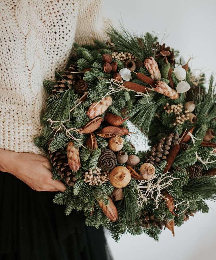 Uma pessoa segurando uma guirlanda de Natal feita com galhos de pinheiro, pinhas, canela e elementos naturais secos. Ela veste um suéter de tricô creme e uma saia preta. A guirlanda tem um visual rústico e elegante, ideal para decoração.