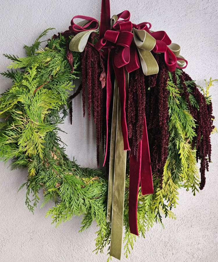 Enfeite de Natal de porta feito com ramos verdes e detalhes de folhagens vermelhas. No topo, laços de veludo vermelho e verde criam um destaque elegante. O fundo é uma parede clara, ressaltando as cores vibrantes e o design festivo do enfeite.