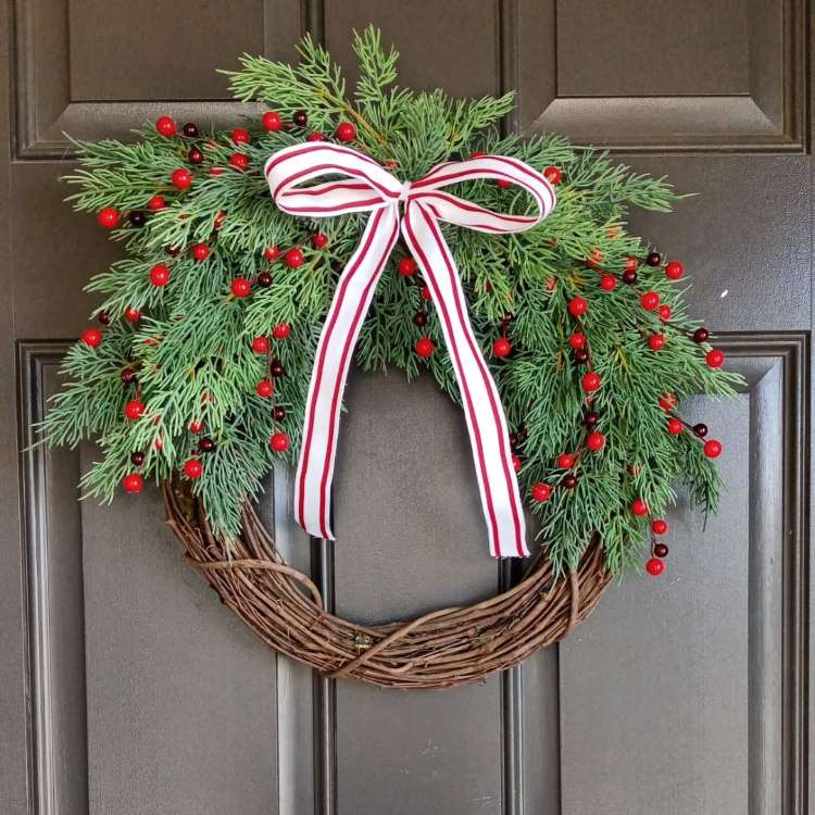 Guirlanda de Natal feita de ramos entrelaçados, adornada com um laço xadrez vermelho no topo. Na base, pinhas, galhos verdes e bagas vermelhas. Pequenos pássaros vermelhos decoram o lado direito, criando um visual festivo e acolhedor.