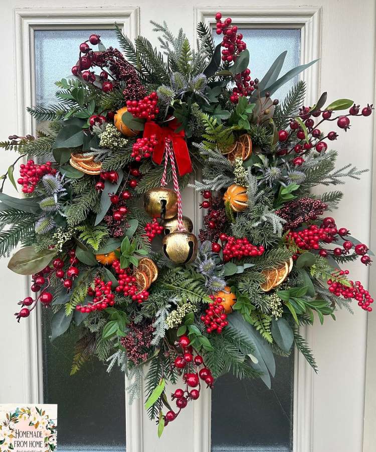 Enfeite de Natal de porta exuberante com ramos verdes e bagas vermelhas. Tem fatias de laranja secas e sinos dourados pendurados. Um laço vermelho vibrante adiciona charme, enquanto folhagens variadas criam um visual festivo e natural em uma porta branca.