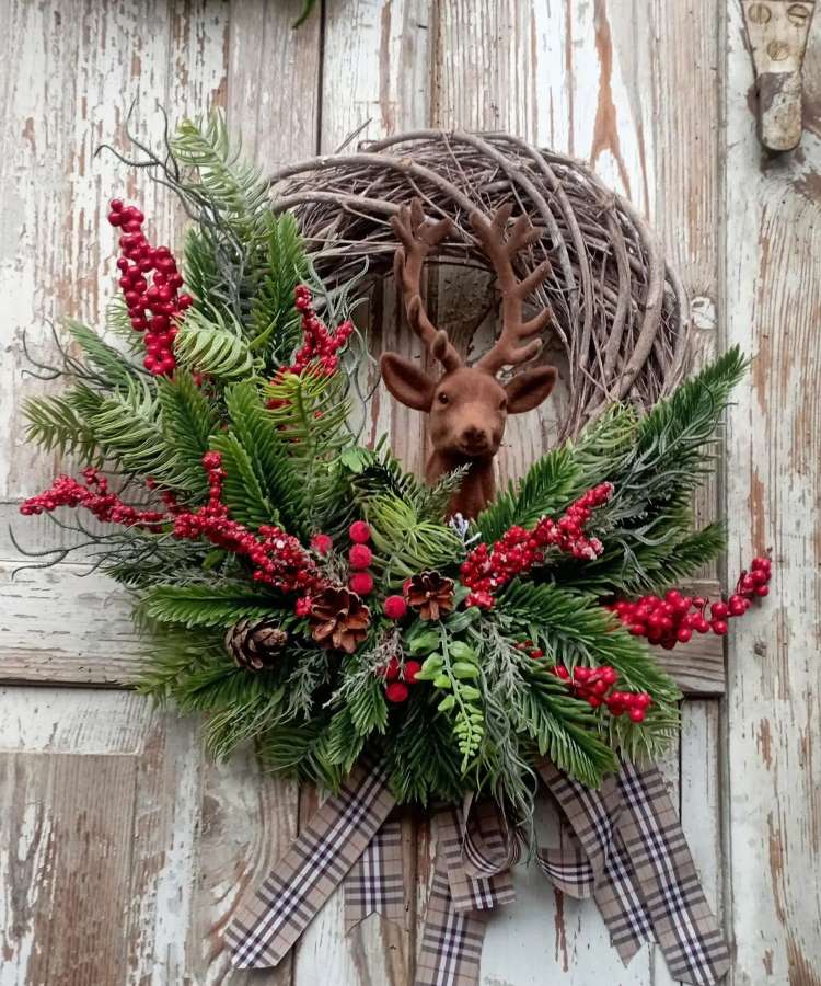 Guirlanda de Natal adornada com folhagens verdes, frutas vermelhas, pinhas e laço xadrez. No centro, destaca-se a cabeça de um cervo. Fundo de madeira envelhecida complementa o visual rústico, criando uma decoração charmosa e festiva para portas e paredes.