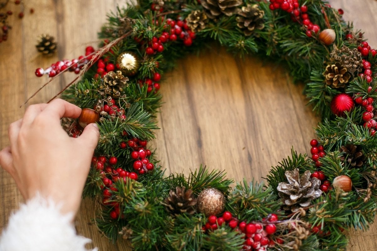 Enfeite de Natal de porta feito com galhos verdes, decorado com pinhas, bagas vermelhas e bolas douradas. Uma mão ajusta um detalhe na composição, sobre uma superfície de madeira clara, criando um toque caseiro e natural na decoração natalina.