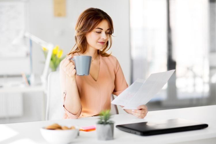 mulher com xícara, segurando papéis, sentada em mesa com notebook e biscoitos