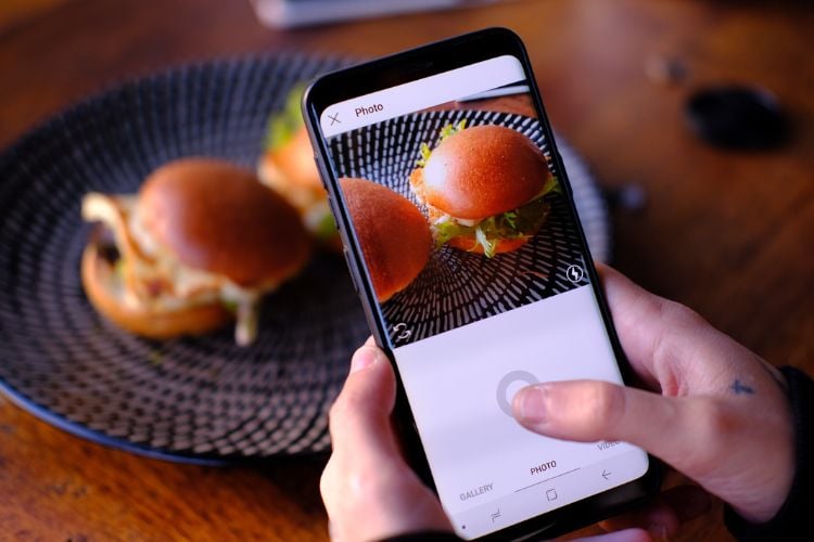 A imagem mostra uma influenciadora tirando uma foto de dois hambúrgueres em um prato preto com detalhes circulares. Ela usa um smartphone, capturando a refeição de cima. A cena transmite um ar casual e gastronômico, com foco na comida.