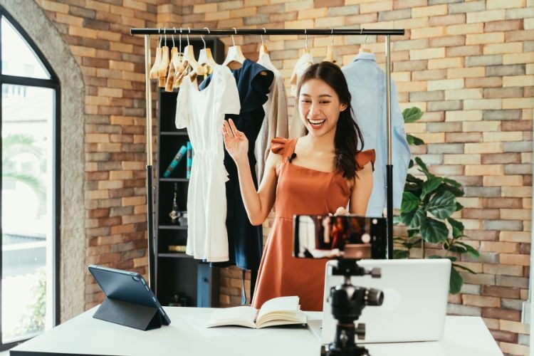 A imagem mostra uma mulher sorridente vestindo um vestido laranja em frente a um celular em um tripé, realizando uma transmissão ao vivo ou gravação. Ao fundo, há roupas penduradas e diversos equipamentos como laptop e tablet.