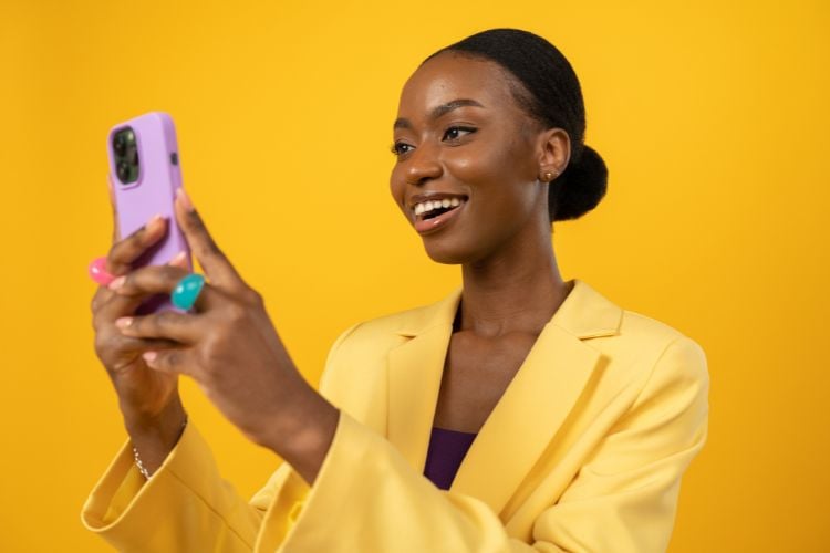 A imagem mostra uma mulher sorridente vestindo um blazer amarelo, segurando um smartphone lilás. Ela tem cabelo preso em um coque e usa anéis grandes e coloridos. O fundo é amarelo, criando uma atmosfera vibrante e alegre.