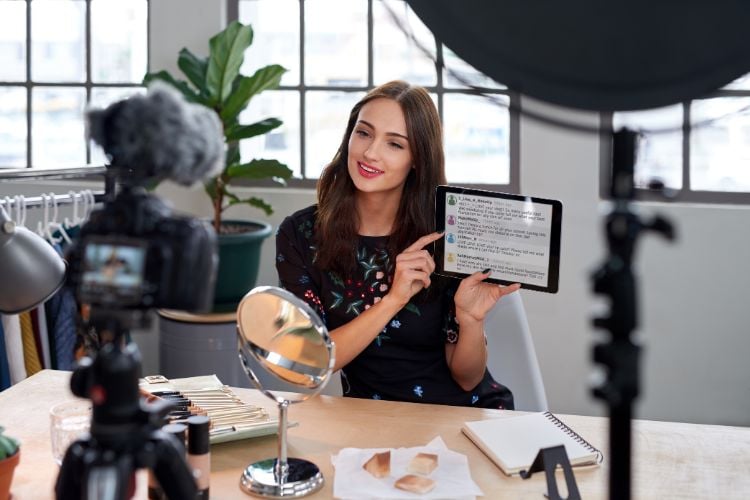 A influencer digital está em um estúdio iluminado, sorrindo enquanto segura um tablet e aponta para a tela. À sua frente, há uma câmera, espelhos, blocos de anotações e maquiagem. Ao fundo, plantas e roupas complementam o ambiente criativo e profissional.