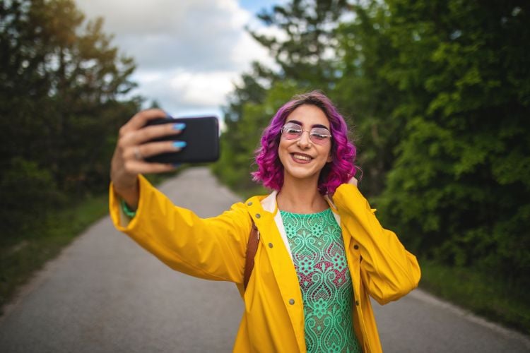 A imagem retrata uma influencer digital sorridente com cabelo roxo e óculos, tirando uma selfie em um caminho ao ar livre. Ela veste um casaco amarelo vibrante sobre uma blusa verde. Ao fundo, há árvores verdes sob um céu parcialmente nublado.