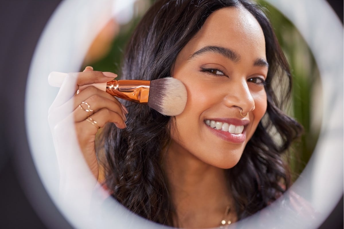 A imagem mostra uma mulher sorrindo enquanto aplica blush com um pincel grande no rosto. Ela tem cabelo ondulado, brincos pequenos e um piercing no nariz. O reflexo no espelho circular cria um enquadramento suave. O ambiente é iluminado.