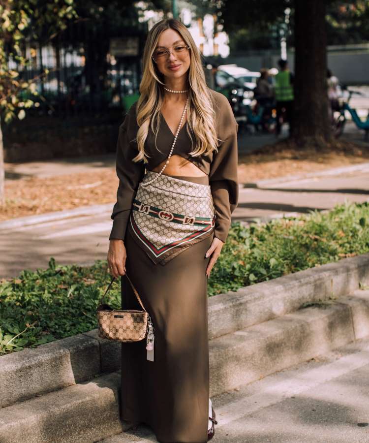 A mulher está em um ambiente ao ar livre, usando um vestido marrom longo com recortes. Ela adiciona um lenço estampado na cintura e segura uma bolsa pequena. Seus cabelos loiros estão soltos, e ela usa um colar de pérolas, criando um visual sofisticado.