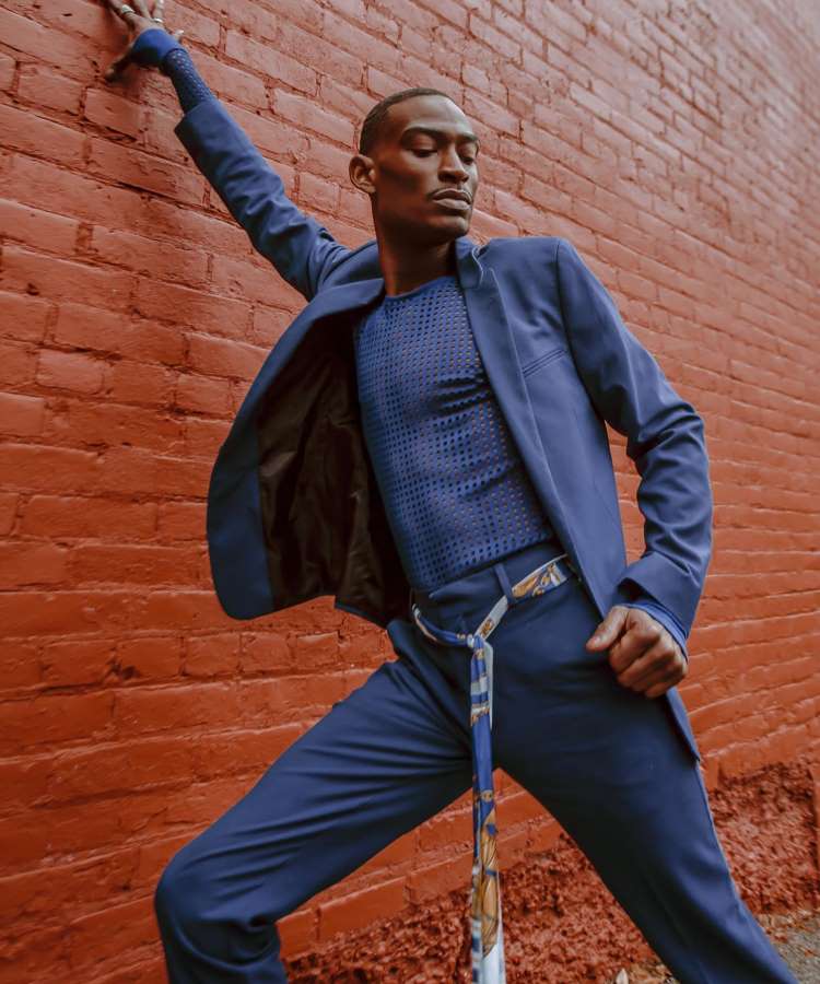 O homem está posando contra uma parede de tijolos vermelhos, usando um terno azul vibrante e uma camiseta texturizada. Ele segura a parede com uma mão, enquanto o lenço estampado amarrado na cintura adiciona um toque de cor ao visual sofisticado.