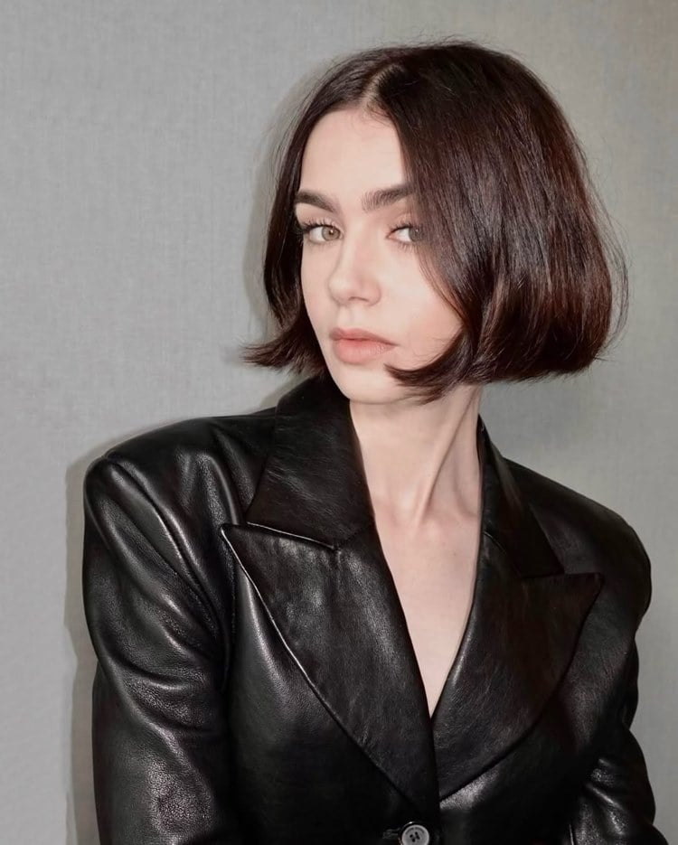 Lily Collins com cabelo curto e liso olha diretamente para a câmera. Ela veste um blazer preto de couro. Sua expressão é séria e confiante, destacando um estilo elegante e moderno. O fundo neutro realça sua presença marcante.