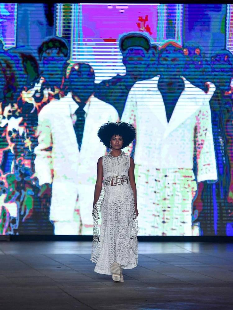 Mulher de pele negra desfilando com vestido branco. Ao fundo, telão com imagem de pessoas