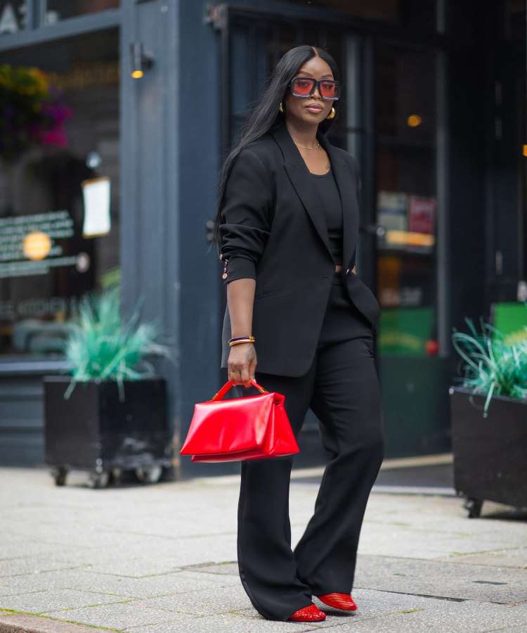 A mulher caminha confiante em uma rua urbana, vestindo um elegante terno preto com calças largas. Ela segura uma bolsa vermelha vibrante que combina com seus sapatos e óculos de sol. Este é um dos "Looks para festa de empresa" que destacam estilo e ousadia.