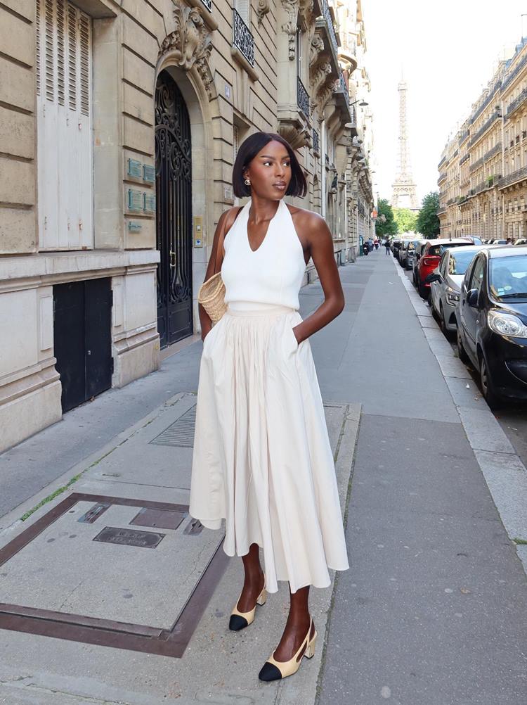 Uma mulher elegante caminha por uma rua parisiense, vestindo um conjunto branco sofisticado. Ao fundo, a Torre Eiffel se destaca, cercada por edifícios clássicos e carros estacionados. O cenário exala charme e estilo urbano, típico de Paris.