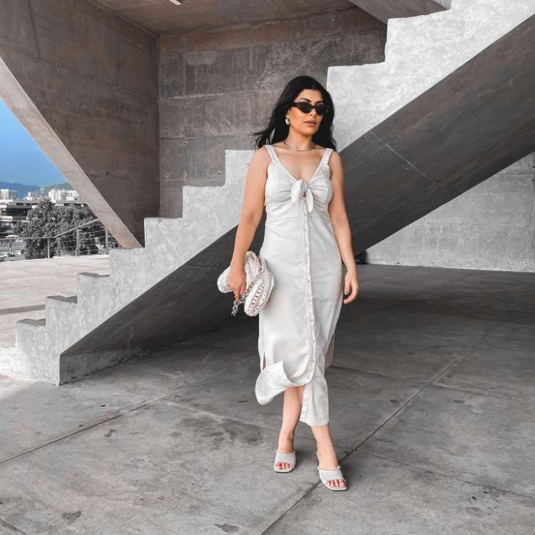 Uma mulher usa um vestido branco elegante e óculos de sol, caminhando por um espaço moderno com escadarias de concreto. Ela segura uma bolsa texturizada e está em um ambiente urbano, com prédios e montanhas visíveis ao fundo, sob um céu azul.