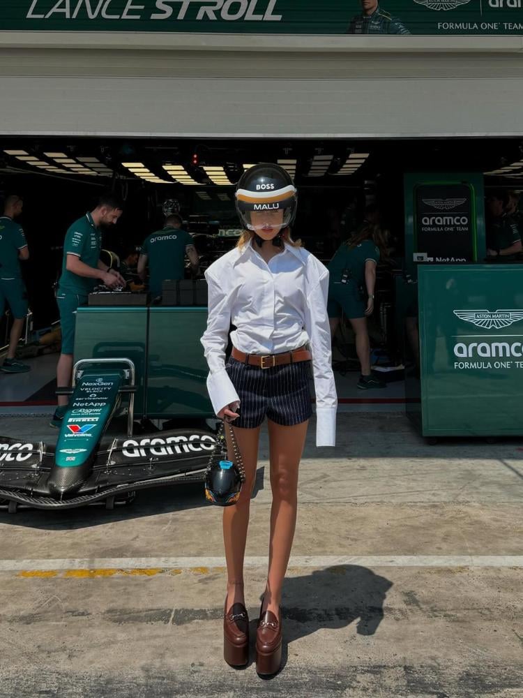 Malu Borges em frente ao box da Aston Martin usando camisa branca de ombro largo, capacete personalizado "Malu" e "Boss", bolsa de capacete, shorts listrado e mocassim marrom de plataforma