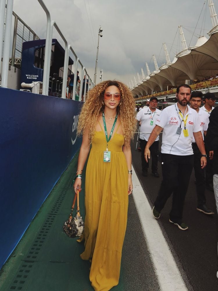 Julia Rodrigues no paddock usando vestido longo amarelo, óculos de sol e bolsa marrom