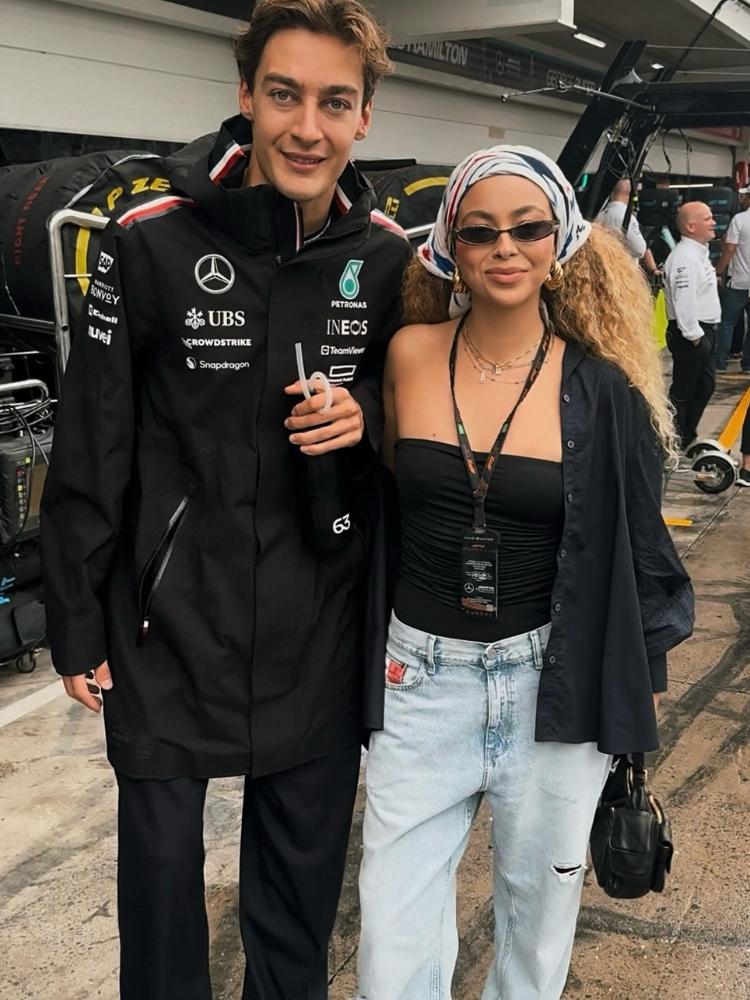 George Russel e Julia Rodrigues abraçados. Ele usa roupa da Mercedes e ela usa body sem alça preto, camisa preta por cima, calça jeans e lenço no cabelo