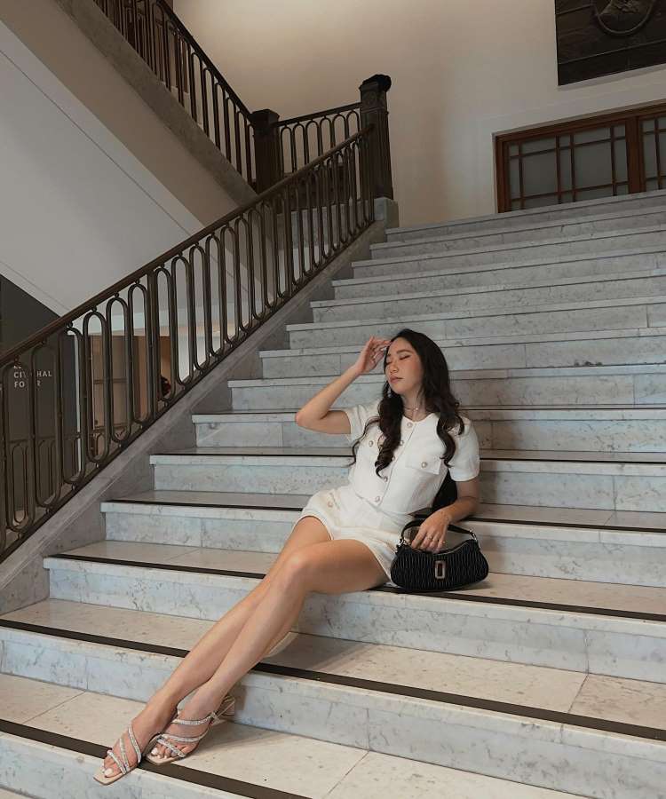 Mulher sentada em escada de mármore, usando vestido branco curto e sandálias de salto. Bolsa preta ao lado. Ambiente interno elegante, com corrimão de metal ornamental. Olhar pensativo, pose relaxada.