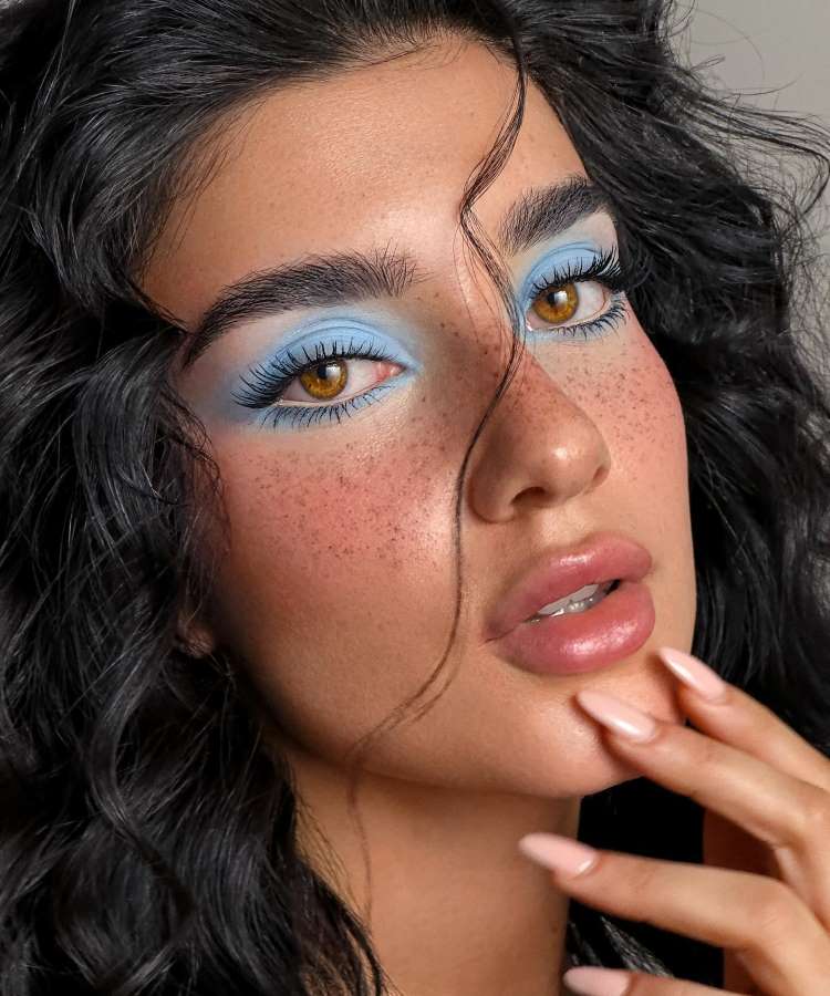 A imagem mostra uma mulher com maquiagem vibrante. Seus olhos têm sombra azul clara, realçando os olhos castanhos e cílios longos. A pele é adornada com sardas sutis, e seus lábios são rosados. Cabelos pretos e ondulados emolduram o rosto expressivo.