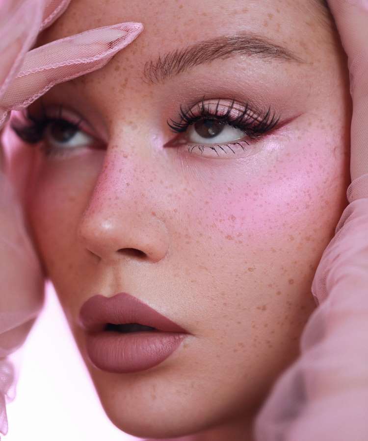 A imagem apresenta um close-up de uma mulher com maquiagem delicada. Seus olhos são realçados por cílios longos e delineado fino, com sombra rosada. A pele exibe sardas suaves, e suas bochechas têm um blush rosa. Os lábios são cor de malva. Ela toca o rosto com luvas de tecido rosa transparente.