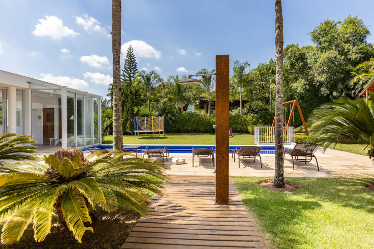 A imagem retrata um jardim exuberante com uma piscina azul brilhante cercada por espreguiçadeiras. Há palmeiras altas e vegetação ao redor, proporcionando uma atmosfera tropical. Ao fundo, uma moderna estrutura de vidro sugere uma área de convivência. O céu está azul, pontuado por nuvens brancas fofas. A imagem retrata um jardim exuberante com uma piscina azul brilhante cercada por espreguiçadeiras. Há palmeiras altas e vegetação ao redor, proporcionando uma atmosfera tropical. Ao fundo, uma moderna estrutura de vidro sugere uma área de convivência. O céu está azul, pontuado por nuvens brancas fofas.