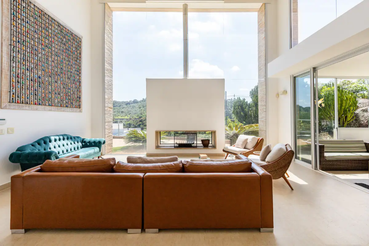 A foto mostra uma sala de estar espaçosa com um sofá marrom e um sofá azul vibrante. A lareira moderna é central, e grandes janelas de vidro oferecem vistas para a vegetação externa. Há uma obra de arte colorida na parede e poltronas confortáveis, criando um ambiente sofisticado e acolhedor.