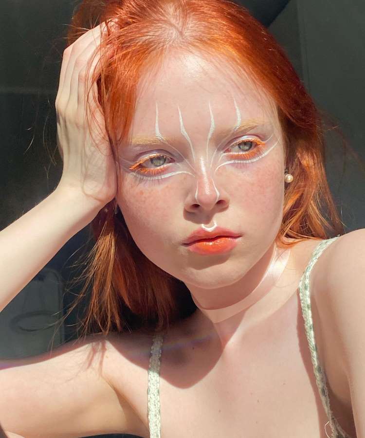 A imagem mostra uma jovem de cabelo ruivo, com maquiagem artística em branco e laranja ao redor dos olhos. Sua expressão é contemplativa, com luz destacando a pele clara. Ela usa uma roupa leve e apoia a mão na cabeça.