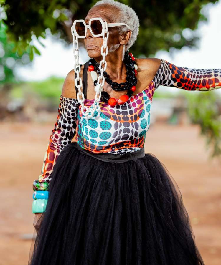 Margaret Chola exibe um estilo vibrante com óculos de sol quadrados, correntes brancas e uma blusa estampada colorida. Ela usa uma saia preta volumosa e acessórios grandes, posando ao ar livre, irradiando confiança e originalidade em um ambiente natural.