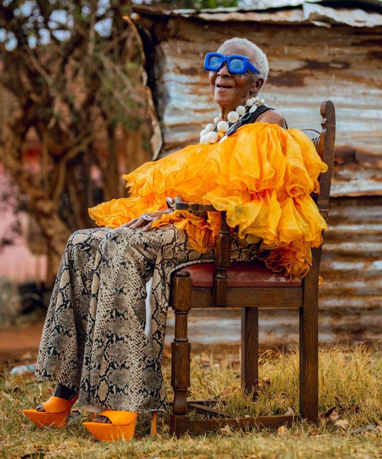 Uma senhora está sentada em uma cadeira de madeira, usando uma blusa laranja volumosa e calças estampadas de cobra. Seus óculos azuis são grandes e ousados, enquanto colares de pérolas brancas adornam seu pescoço. Sapatos de plataforma laranja completam o visual vibrante.