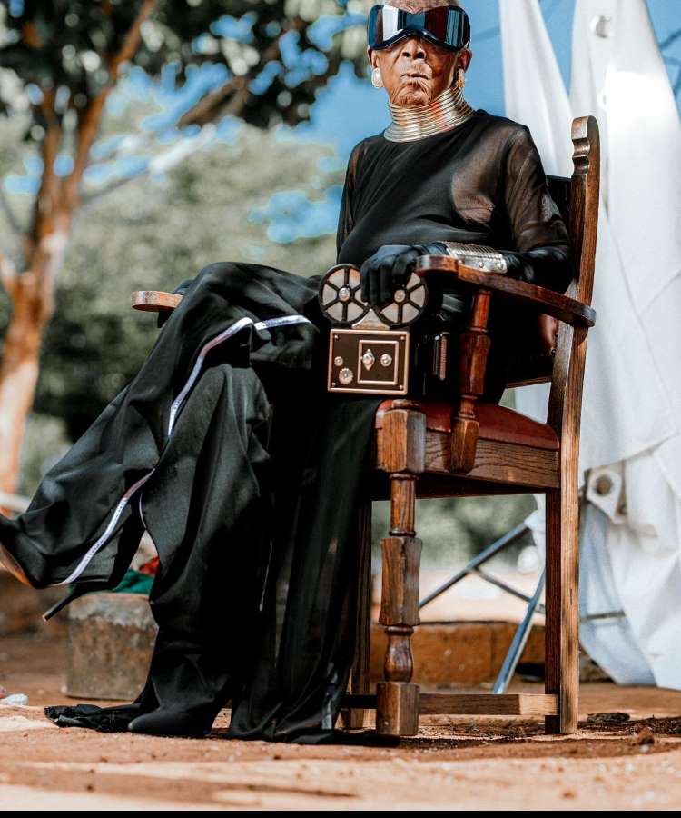 Margaret Chola está sentada em uma cadeira de madeira, usando um elegante vestido preto transparente. Seus grandes óculos de sol e colar dourado chamam atenção. Com luvas pretas e um acessório futurista em mãos, ela exala confiança em um ambiente externo.