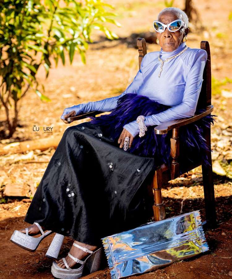 Margaret Chola está sentada em uma cadeira de madeira ao ar livre, usando uma blusa azul brilhante e uma saia preta com detalhes. Seus óculos de sol são chamativos, e ela complementa o visual com sapatos de plataforma prateados e uma bolsa metálica.