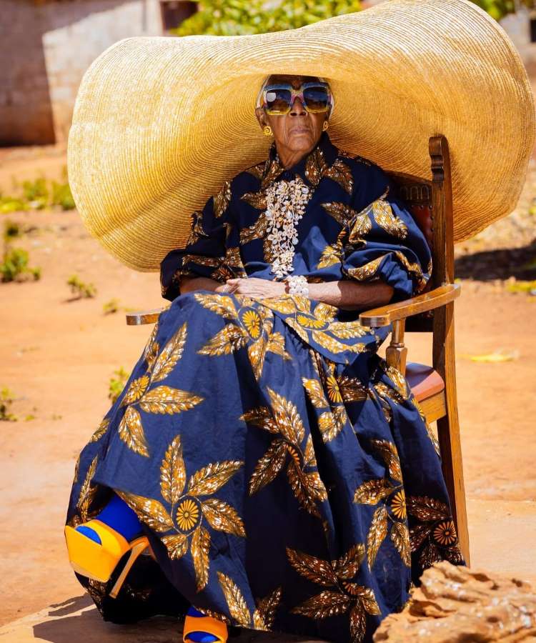 Uma senhora em uma cadeira ao ar livre usa um vestido azul com estampas douradas, acompanhado por um chapéu de palha enorme. Seus óculos são grandes e elegantes, e ela usa colares volumosos. Sapatos de plataforma azul e laranja completam o visual distinto.