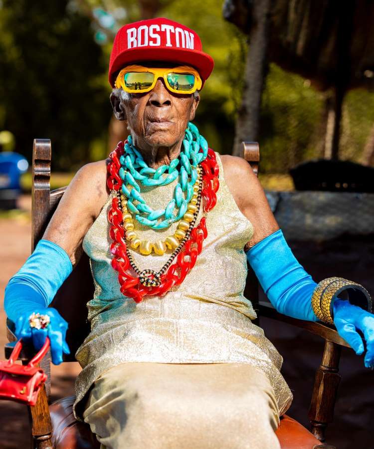 Margaret Chola está sentada em uma cadeira de madeira, usando um vestido dourado. Ela exibe um boné vermelho com "Boston", óculos amarelos e colares grandes coloridos. Suas luvas azuis e pulseiras douradas completam o visual ousado e vibrante em um ambiente ensolarado.