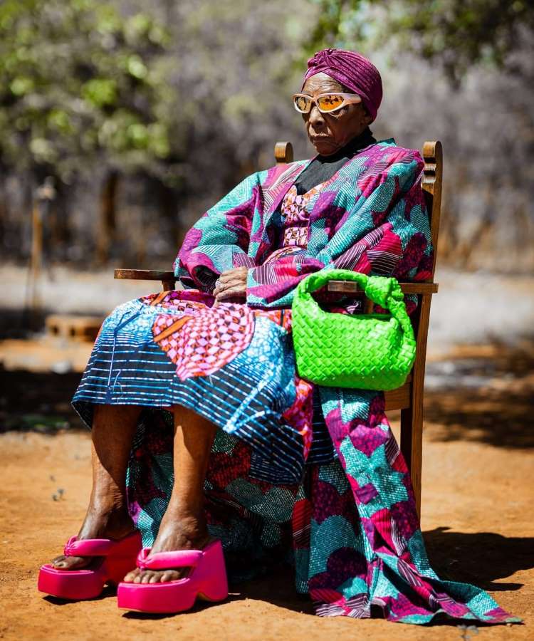 Margaret Chola está sentada ao ar livre, usando um vestido colorido com padrões vibrantes. Ela tem um turbante roxo e óculos de sol elegantes. Sua bolsa verde e sapatos rosa chamam atenção, refletindo seu estilo audacioso e inspirador em um ambiente natural.