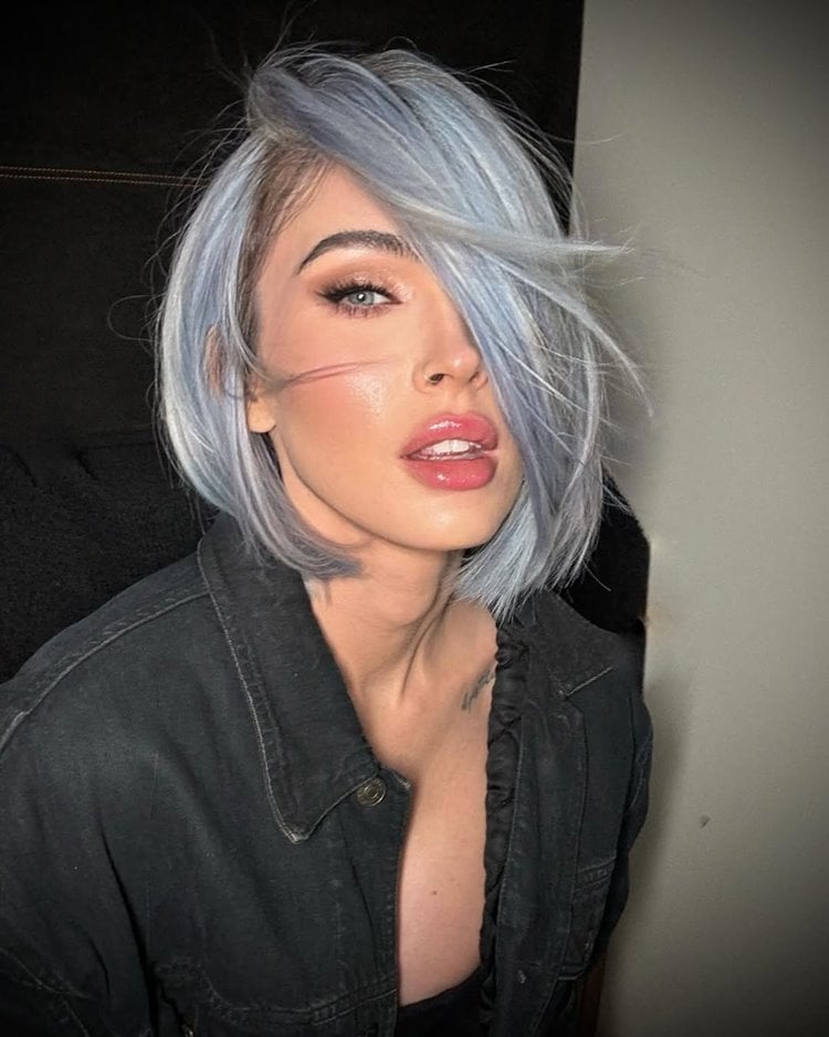 A imagem mostra Megan Fox com corte bob azul-prateado, penteado de lado. Ela tem maquiagem marcante com delineado forte e batom rosa. Veste uma jaqueta escura. A expressão é confiante e sedutora, com iluminação suave destacando suas características faciais.