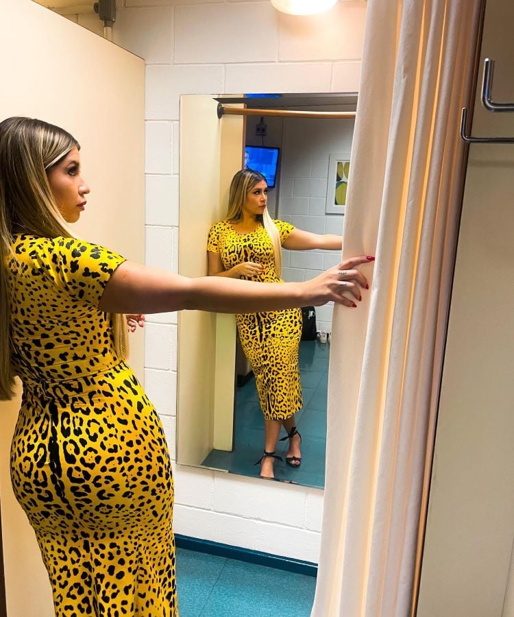 Marília Mendonça em provador, usando vestido amarelo estampado de oncinha, observa-se no espelho. Ela tem cabelos longos e loiros. O chão, revestido de carpete azul, contrasta com as paredes brancas de tijolos aparentes. Atrás dela, uma cortina bege está aberta, revelando o ambiente de loja.