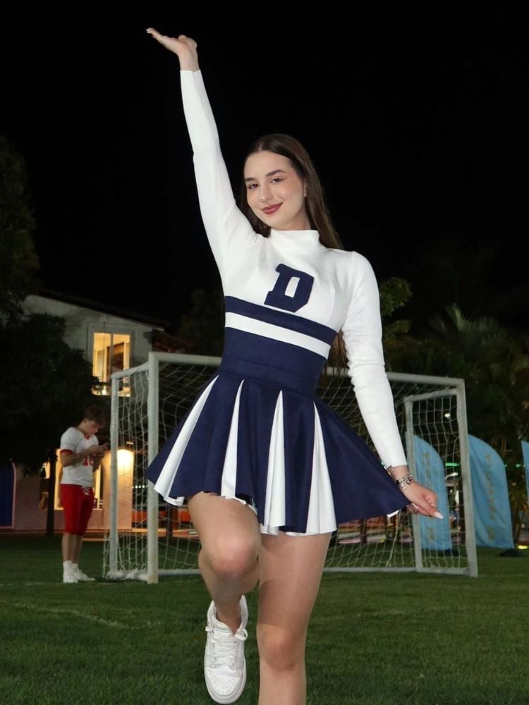 Duda Rubert em campo de futebol usando vestido de líder de torcida branco e azul com o D no colo 