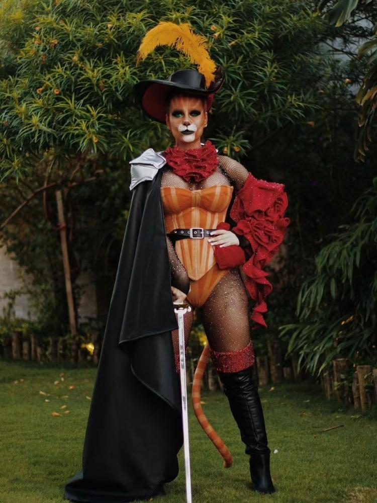 Mulher de pele morena fantasiada de gato de botas, com maquiagem, chapéu, corset laranja, meia-calça, botas pretas e capa preta 