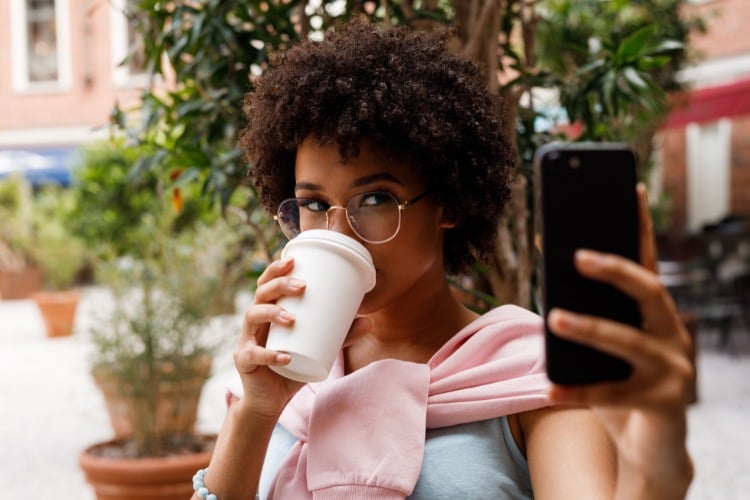 Mulher de pele negra usando óculos, blusa rosa amarrada no pescoço, blusa cinza e tomando café enquanto grava com o celular