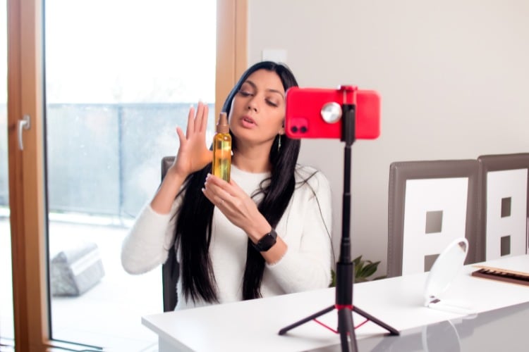 Mulher de pele clara usando roupa branca, sentada à mesa mostrando produto em embalagem marrom para celular vermelho em tripé